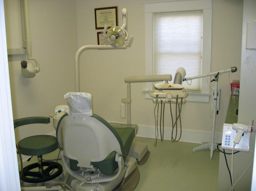 The image shows an interior view of a dental office, featuring a dental chair and equipment, with a sign on the wall and a window with blinds.