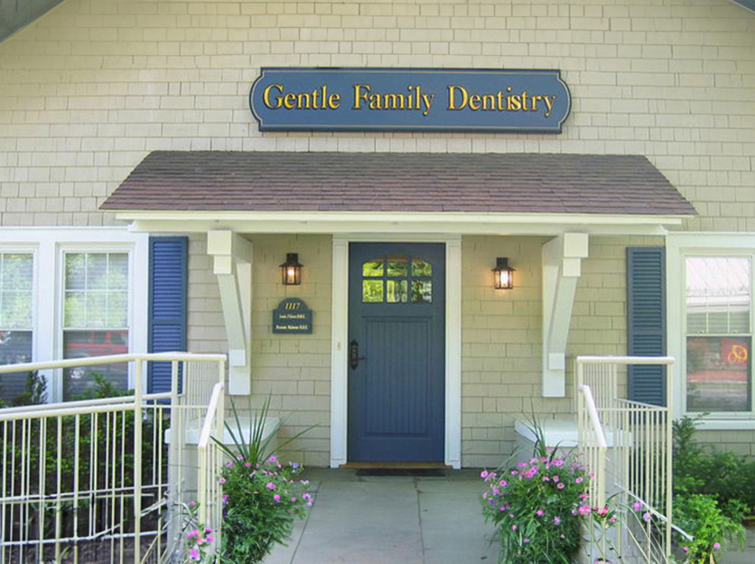 The image is a photograph of a building with the sign  Gentle Family Dentistry  prominently displayed above the entrance. The structure appears to be a single-story commercial building with white siding and a dark blue door, featuring a porch with a railing and steps leading up to it.