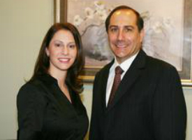 The image shows a man and a woman posing together for a photograph, with the man wearing a suit and tie and the woman in a dark top. They are standing indoors, and there is a framed picture on the wall behind them.