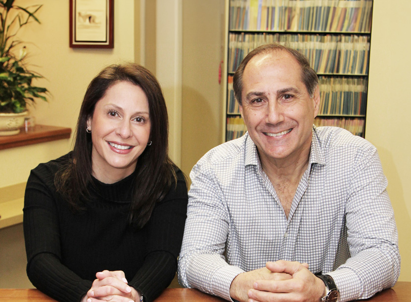 A man and a woman posing together for a photo, both seated at a desk with books behind them.