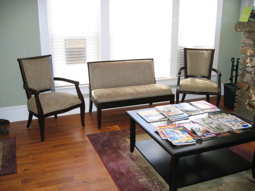 The image depicts a well-lit living room with hardwood floors, a brown sofa, two matching armchairs, a coffee table, and a variety of books and magazines spread out on the table.