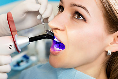 The image shows a woman sitting in a dental chair with a purple mouthpiece inserted into her mouth, receiving dental treatment from a professional wearing a white coat and using a device to scan or examine the teeth.