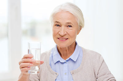 The image shows an elderly woman holding a glass of water, smiling and looking directly at the camera.