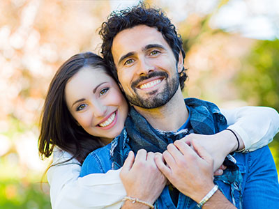 A man and a woman are embracing each other in an outdoor setting, with the man wearing a bandana around his neck.