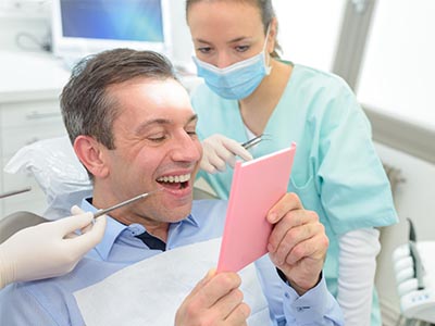 The image shows a man sitting in a dental chair, holding up a pink card with a smile on his face while looking at it. Behind him, there is a female dental professional wearing a surgical mask and standing behind the chair, who appears to be examining the card or showing it to the man. They are both in a dental office setting, as indicated by the presence of dental equipment and the background with a window that lets in natural light.