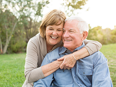 The image shows an elderly couple, a man and a woman, embracing each other outdoors.