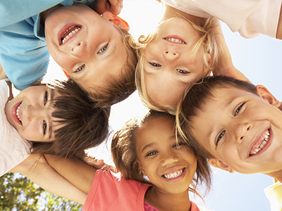 The image shows a group of children, likely in their early to late childhood years, gathered closely together and smiling at the camera. They appear to be outdoors during daylight hours under clear skies.