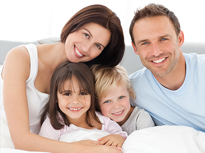 The image features a family of four, including two adults and two children, smiling together in what appears to be a bedroom setting.