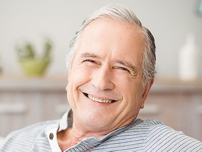 The image depicts an older man with white hair, smiling and looking slightly to the side. He is wearing a blue shirt and has glasses on his face. The background appears to be a room with a light-colored wall and some items that suggest a home setting.