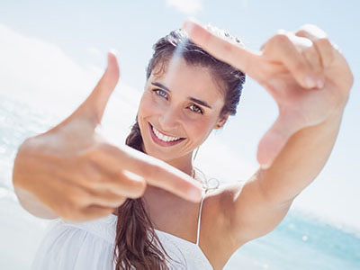 The image is a photograph of a woman outdoors, holding up her hand to frame the camera lens with her fingers, smiling at the camera, and wearing a white top.